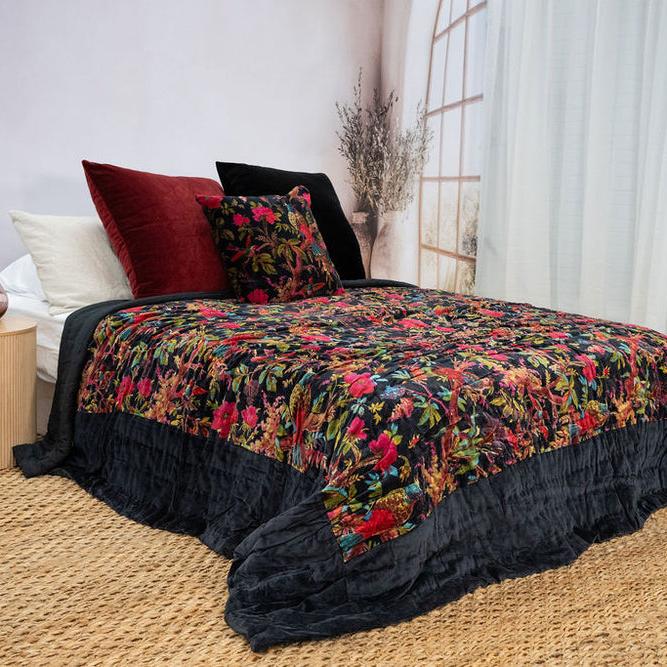 Black velvet quilt with vibrant floral and bird design, red and black pillows, on a bed with a woven rug.