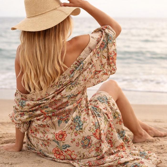 Woman in a beige cotton kimono with colorful floral and bird of paradise print, sitting on a beach.