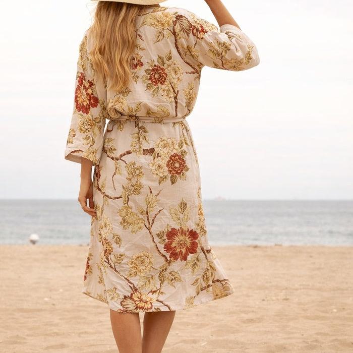 Woman in a cream cotton kimono with red and yellow floral print, tied at waist, on a beach.
