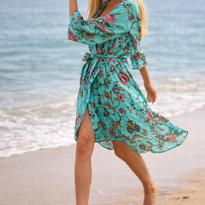 Frida's Vibrance turquoise cotton kimono with colorful floral print, tied at waist, worn on a beach.