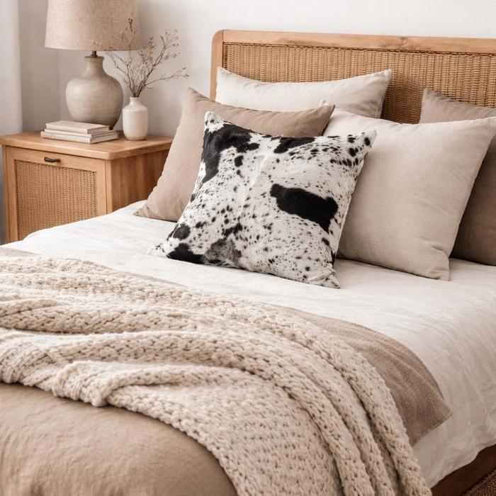 Black and white cowhide cushion cover with natural hair-on texture on a bed with neutral linens.