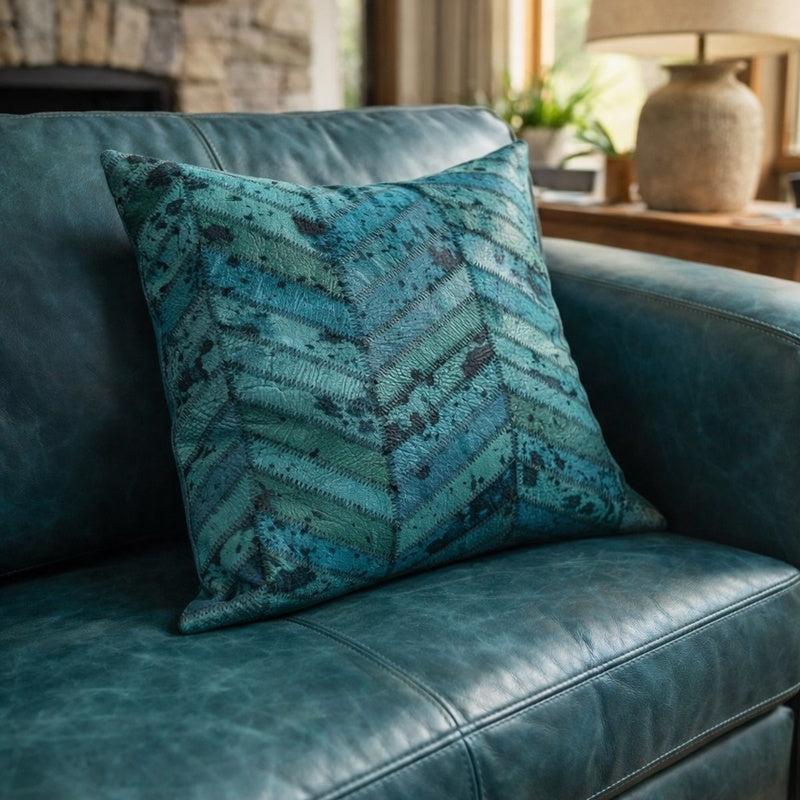 Teal cowhide cushion cover, 40x40cm, with a chevron patchwork design on a blue leather sofa.
