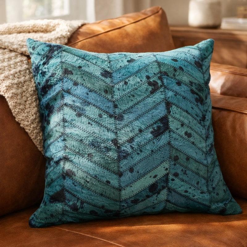 Teal blue patchwork cowhide cushion cover with a chevron pattern on a brown leather sofa.