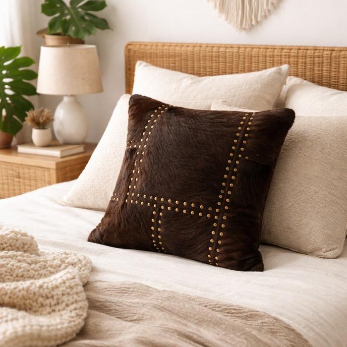 Dark brown cowhide leather cushion cover with gold stud detailing on a bed.