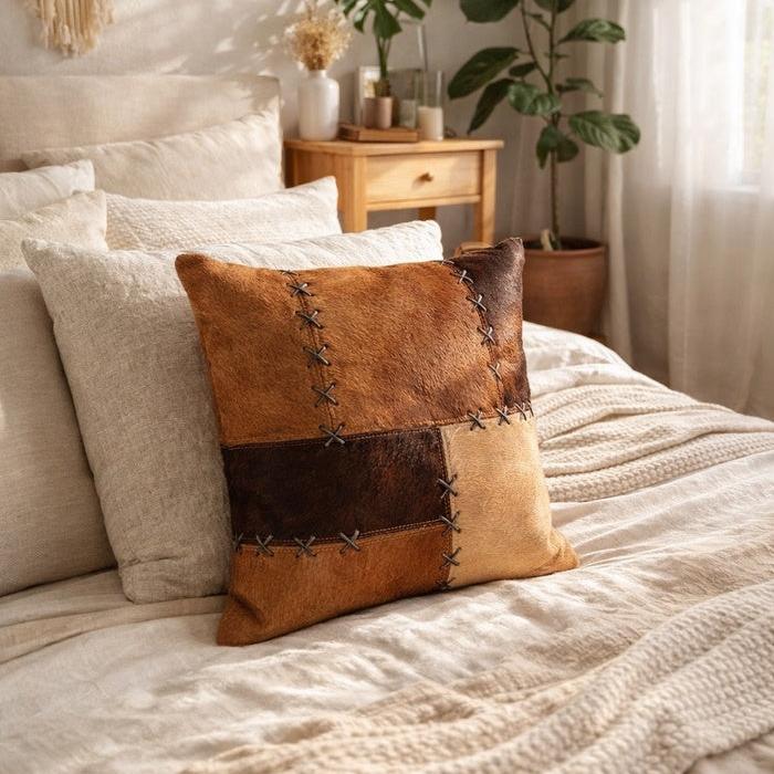Tan & chocolate cowhide leather patchwork cushion cover with cross-stitch lacing on a neutral bed.