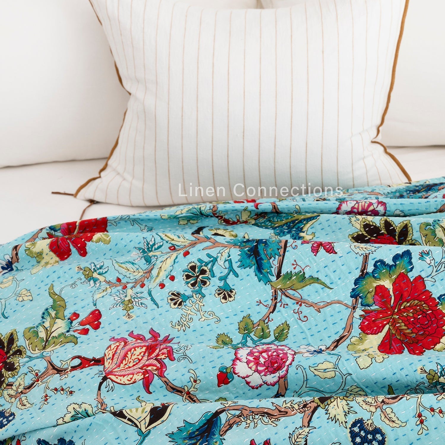 Tropical blue cotton Kantha quilt with vibrant red, pink, and green floral patterns, draped over a white bed.