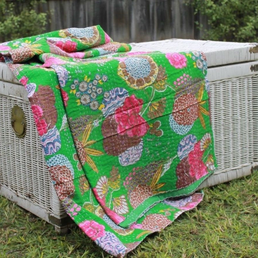 Vibrant green Kantha quilt with pink, brown, and blue floral patterns draped over a white wicker trunk.