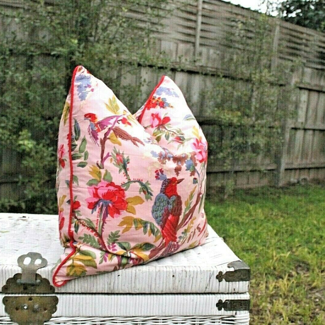 Pink cotton cushion cover with vibrant floral and bird design, red piping, on a white wicker trunk.
