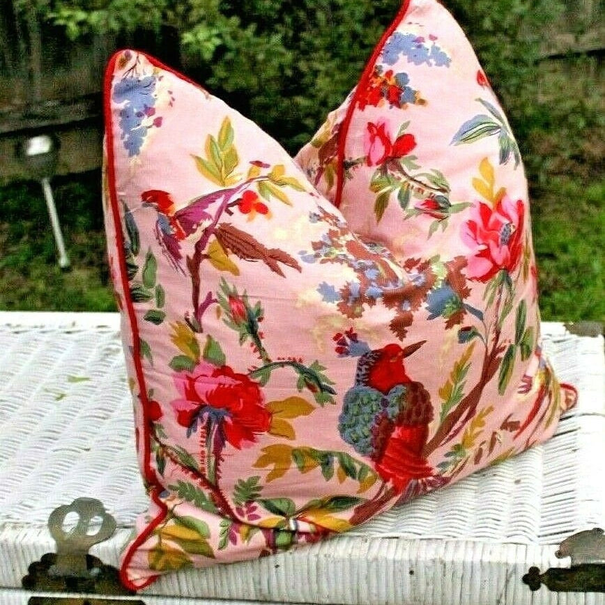 Pink cotton cushion cover with colorful floral and bird print, red piping, on a white wicker surface.