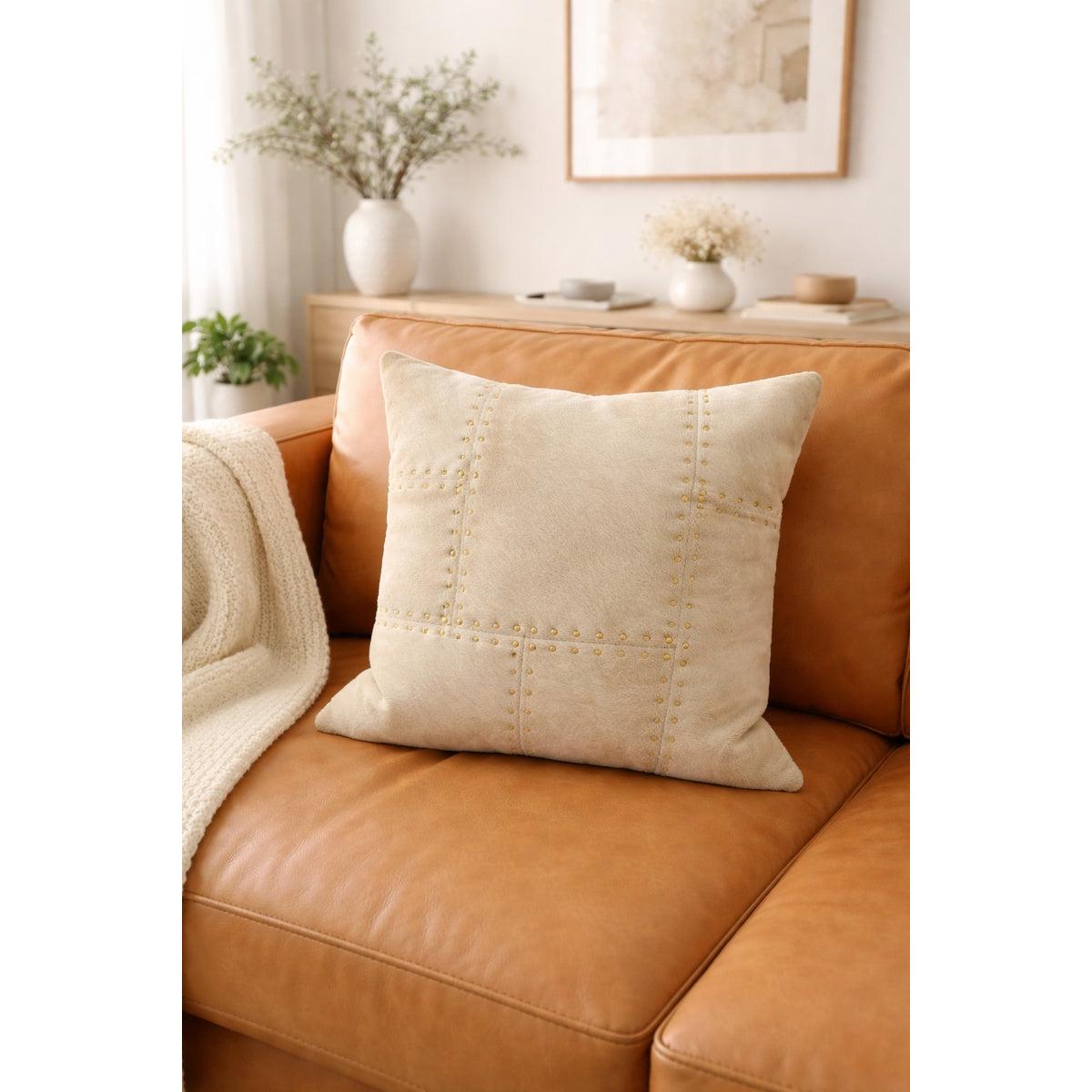 Ivory cowhide leather cushion cover with brass stud detailing on a tan leather sofa.