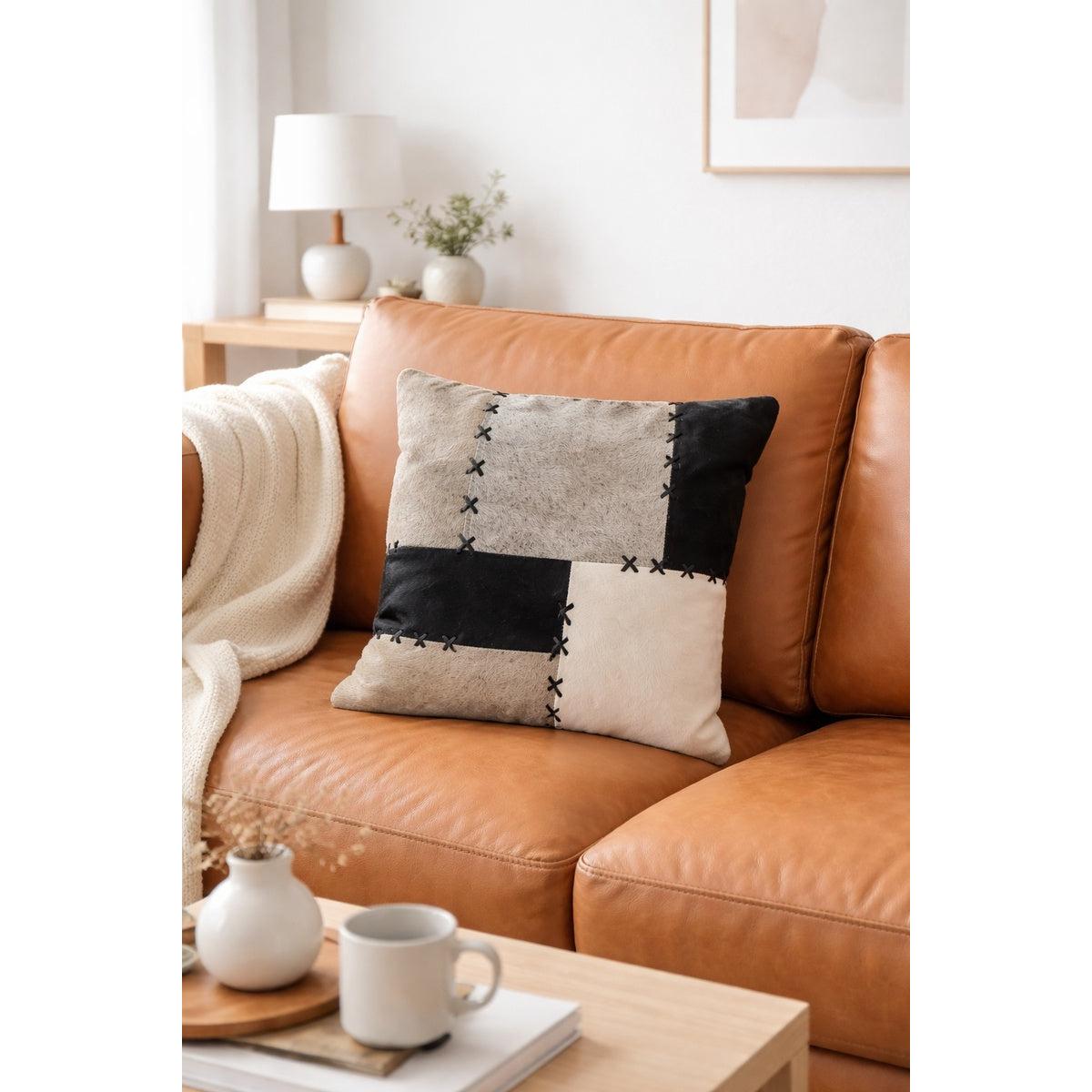 45x45cm black, grey, and cream patchwork cowhide cushion cover with X-stitch detailing on a tan leather sofa.