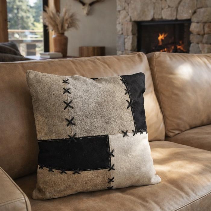 45x45cm black, grey, and cream patchwork cowhide cushion cover with X-stitch lacing on a tan sofa.